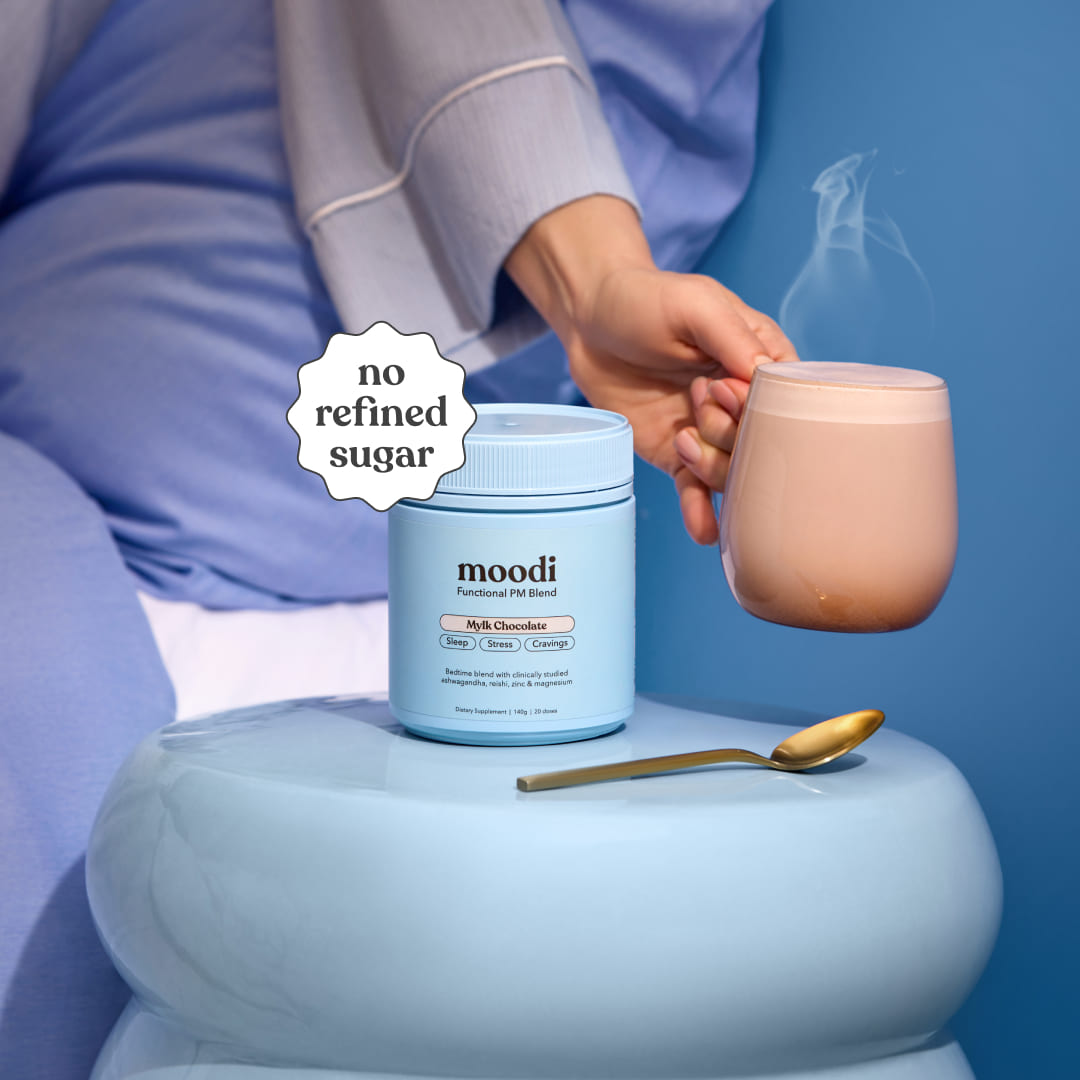 Person holding a steaming mug of Moodi Mylk Chocolate with its tub in the center and a spoon on a blue surface.