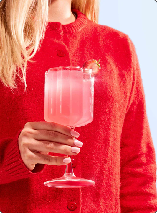 Person wearing a red sweater holding a Moodi Pink Lemonade Pre + Probiotic drink in a glass against a light blue background