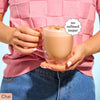 Person wearing a pink shirt and blue jeans holding a mug of Moodi Chai with 'no refined sugar' label