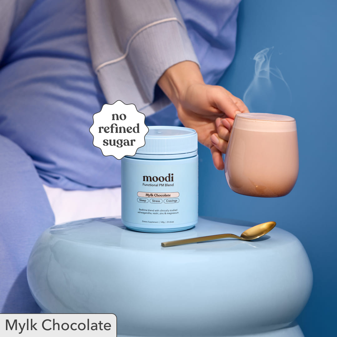 Person holding a steaming mug of Moodi Mylk Chocolate next to its tub with 'no refined sugar' label