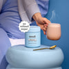 Person holding a steaming mug of Moodi Mylk Chocolate with its tub in the center and a spoon on a blue surface.