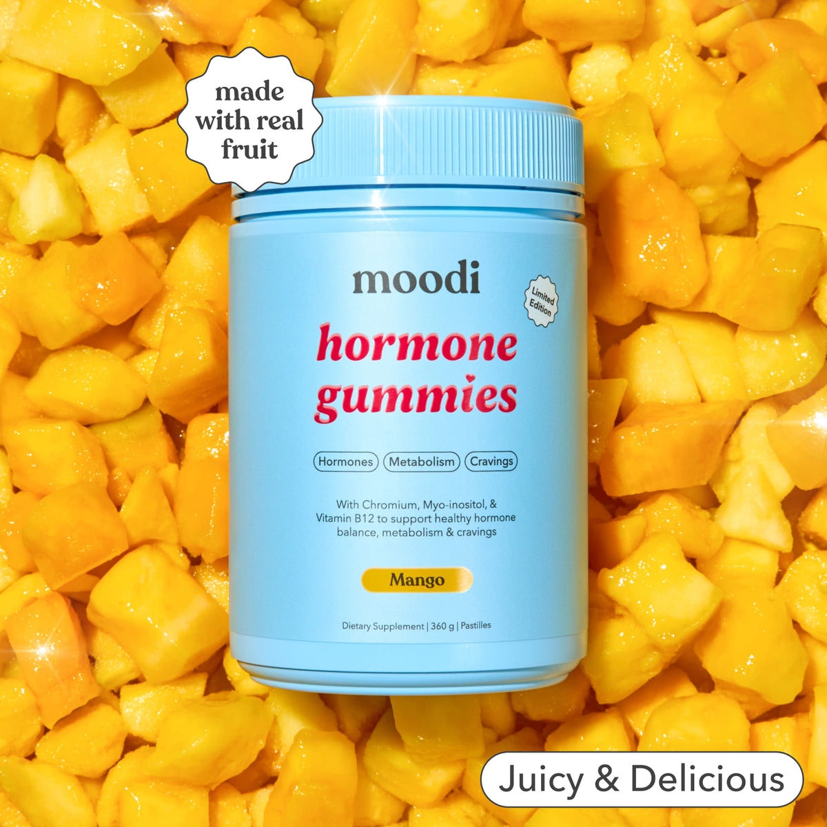 A tub of Moodi Mango Hormone Gummies centered against a background of fresh yellow mango chunks. A white badge in the top left reads 'made with real fruit' and a white banner at the bottom right says 'Juicy & Delicious'.