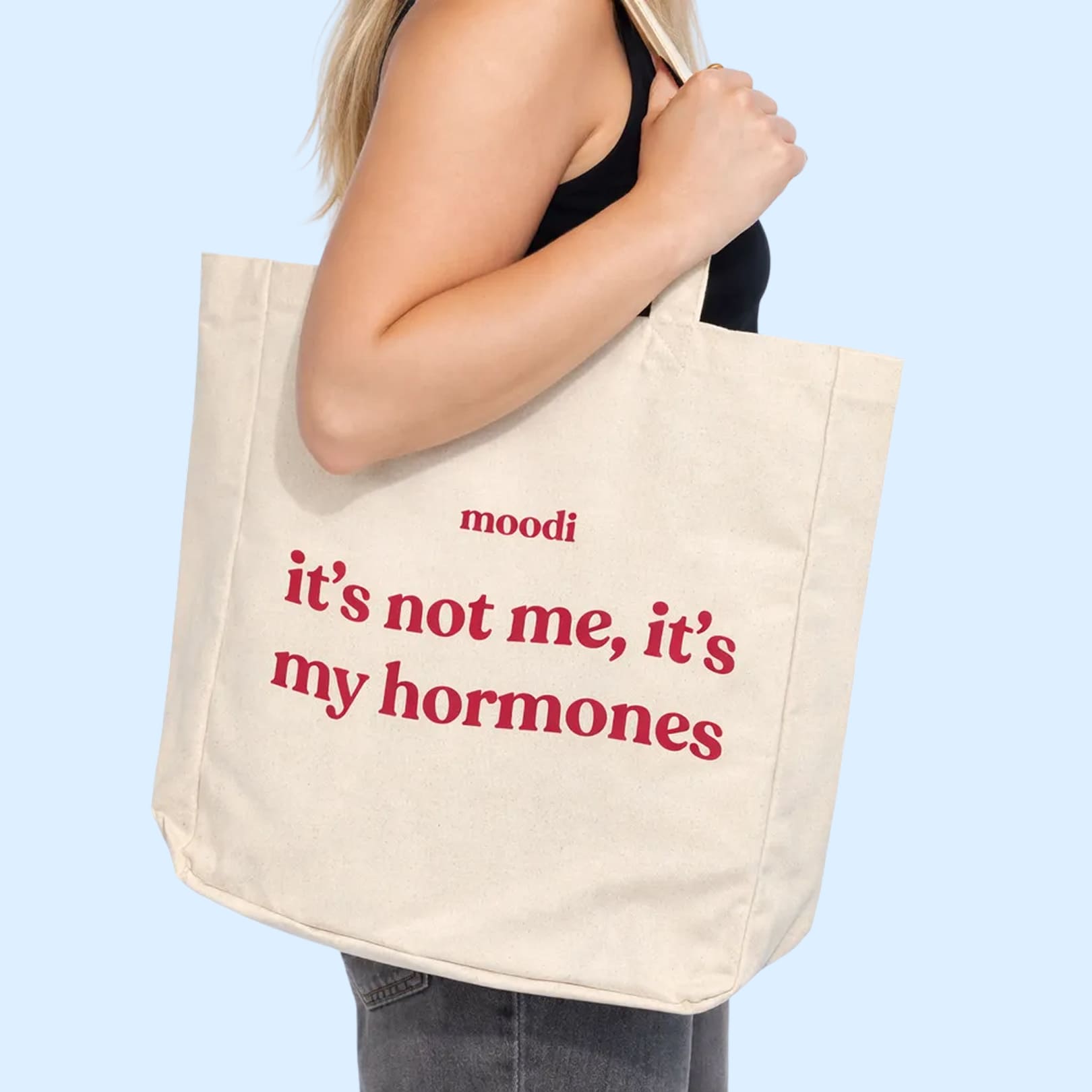 Person holding a tote bag with text 'it's not me, it's my hormones' on a light blue background