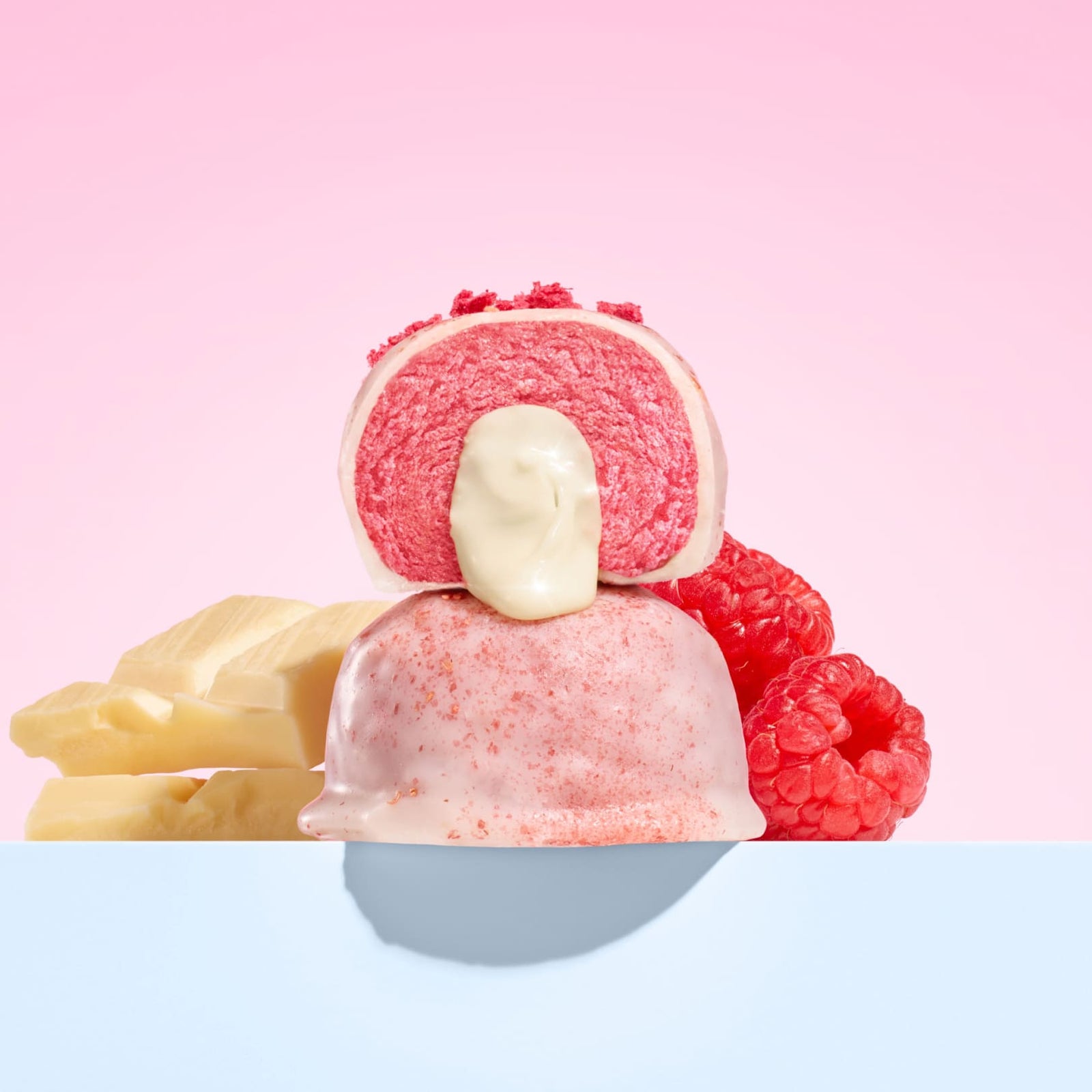 Moodi Raspberry White Choc plant protein ball with white chocolate bars and raspberries on a pink background
