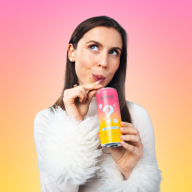 Kate in a white fluffy sweater holds and sips Mystery Flavour Pre + Probiotic Soda against a pink and yellow gradient background